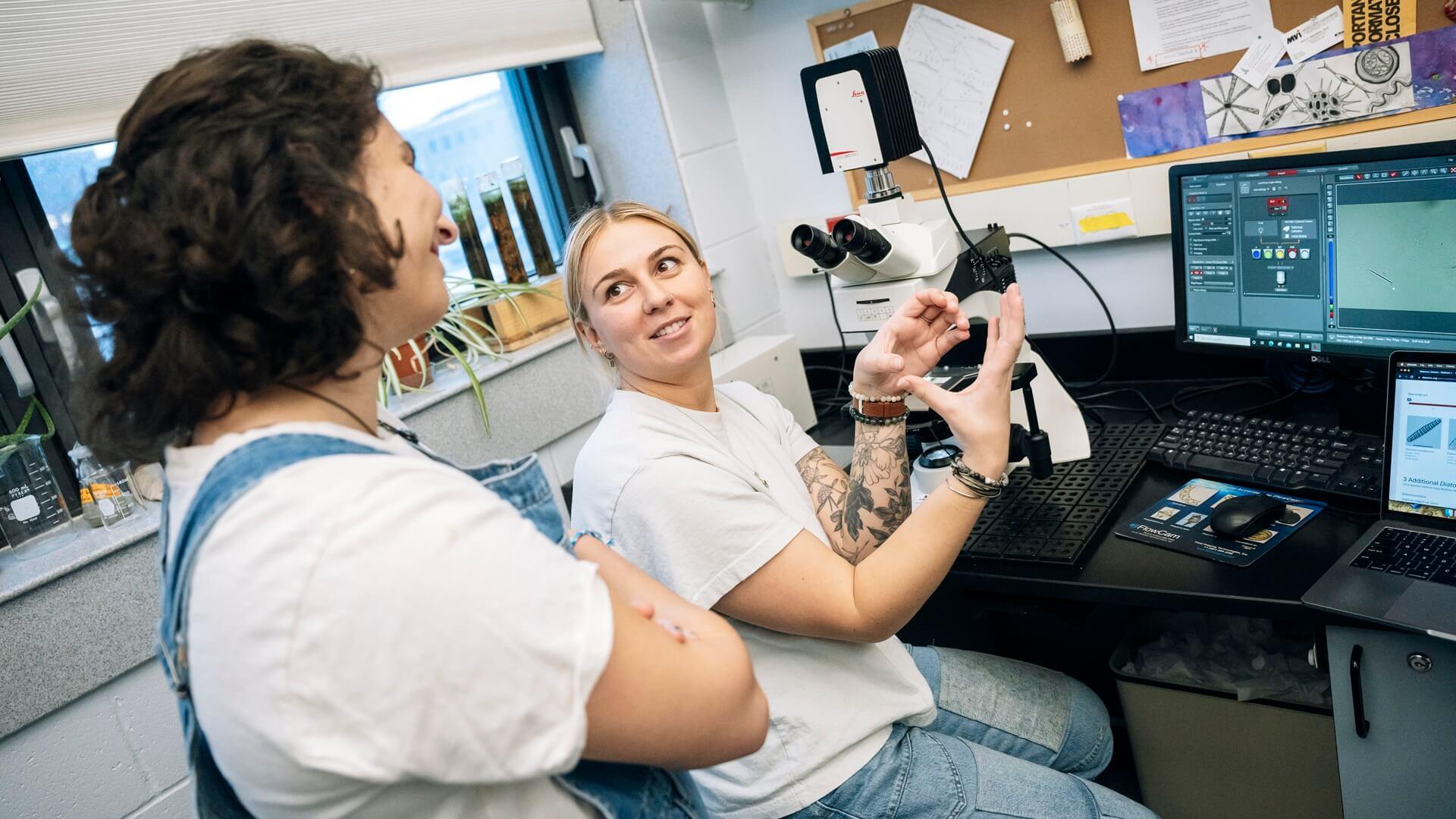 Two students in a lab one is sitting at a microscope and explaining something she is seeing to the other student who is standing and looking at a computer monitor