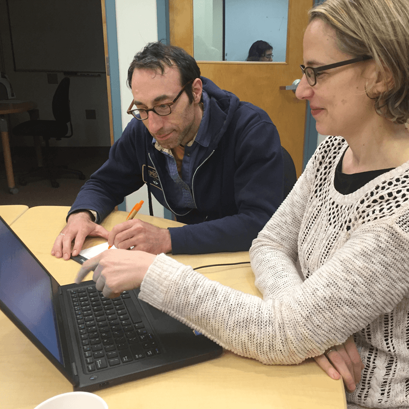 A man (student) and woman (consultant), sit at a writing center table facing a laptop. The student is holding a pen and paper while the consultant points at the laptop screen.