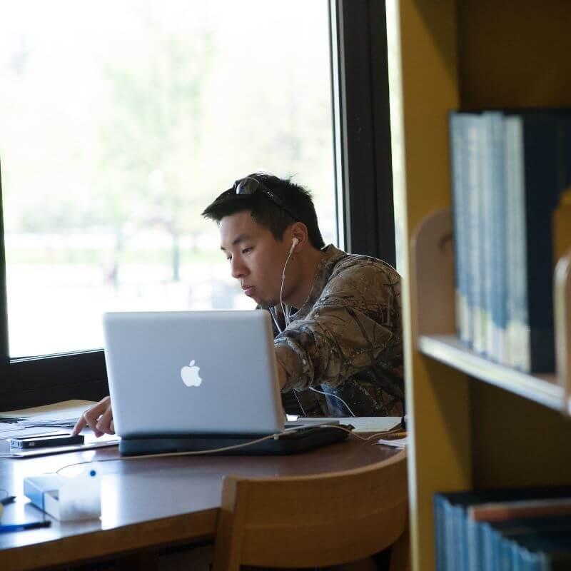 Person sitting at a table in a library with headphones in and laptop open
