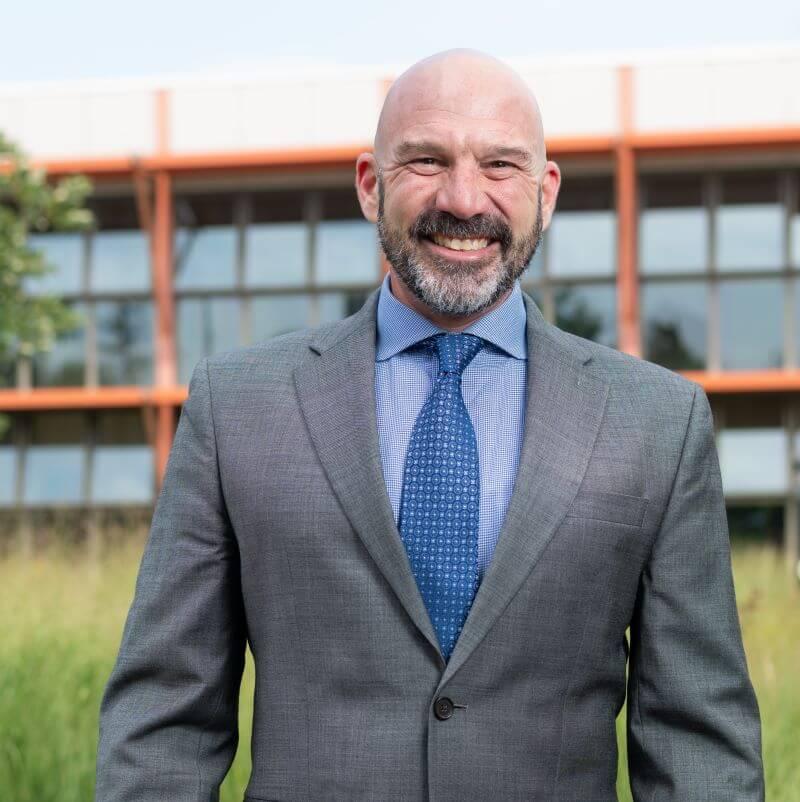 Dean Peter Newman smiling wearing a suit and tie, outside in front of the Aiken Center, the background is blurred