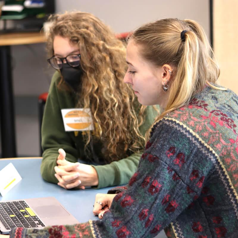 Two women, a writing tutor and a student, sit at a writing center table facing a laptop. One is masked and the other is not.