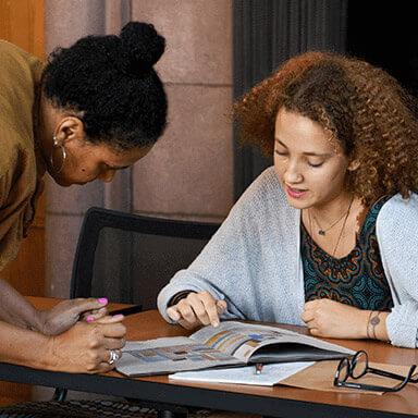 A student examines a book while her professor looks on.