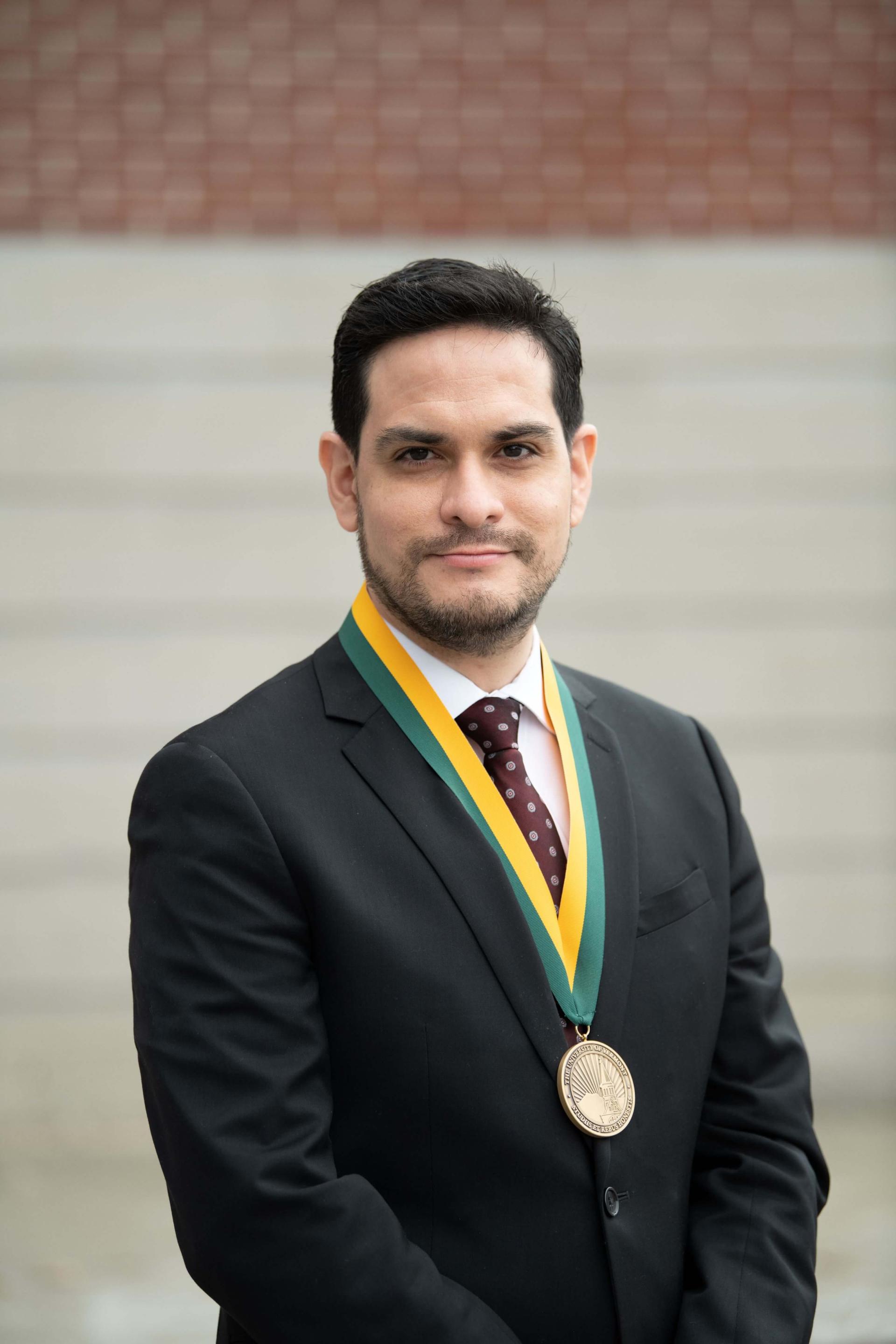 Headshot image of the researcher adorned with an award, posed outdoors in front of a distant building.
