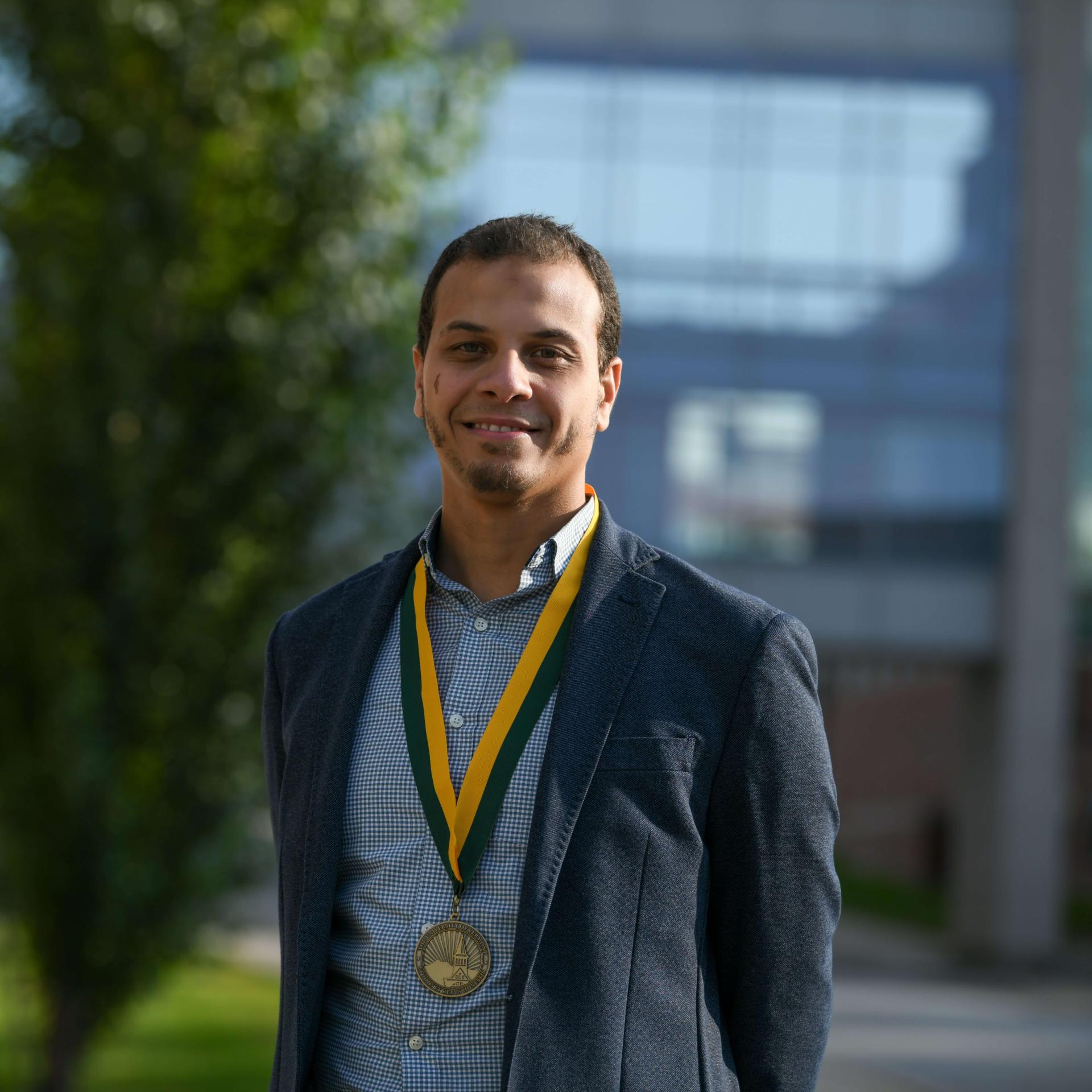 Headshot image of the researcher adorned with an award, posed outdoors in front of a distant building.