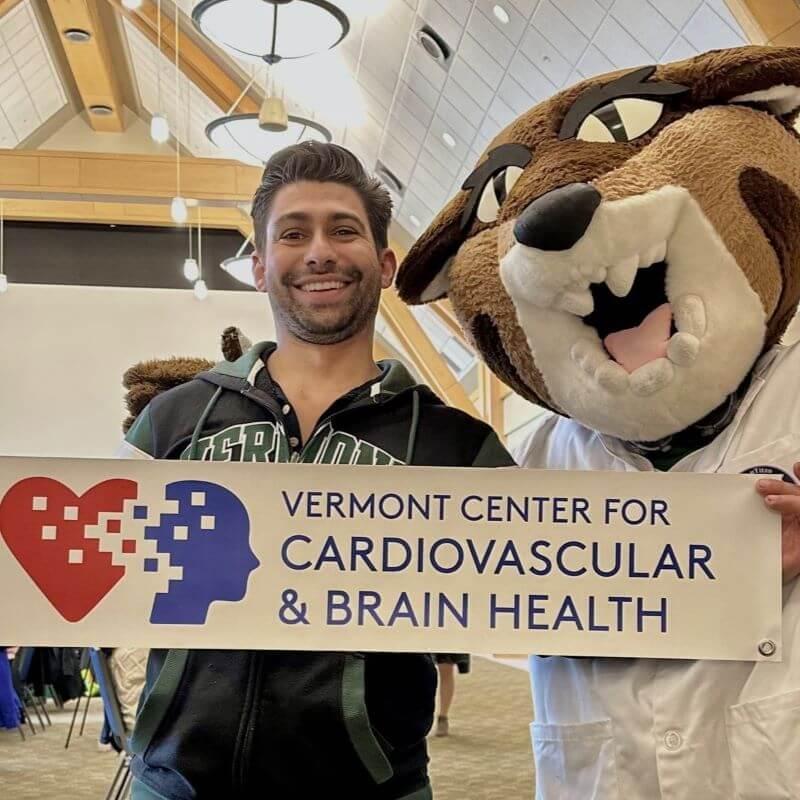 A researcher stands with the University of Vermont's cat mascot, holding a banner that reads "Vermont Center For Cardiovascular & Brain Health" and features the VCCBH logo, which depicts a blended heart and brain image with scattered pixels.