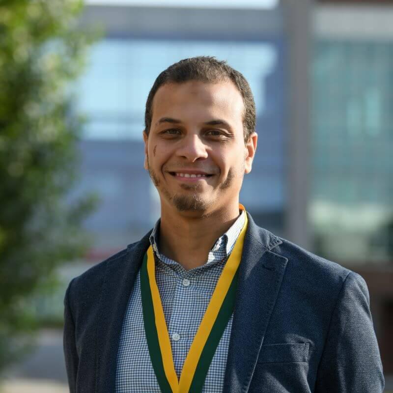 Headshot image of the researcher adorned with an award, posed outdoors in front of a distant building.