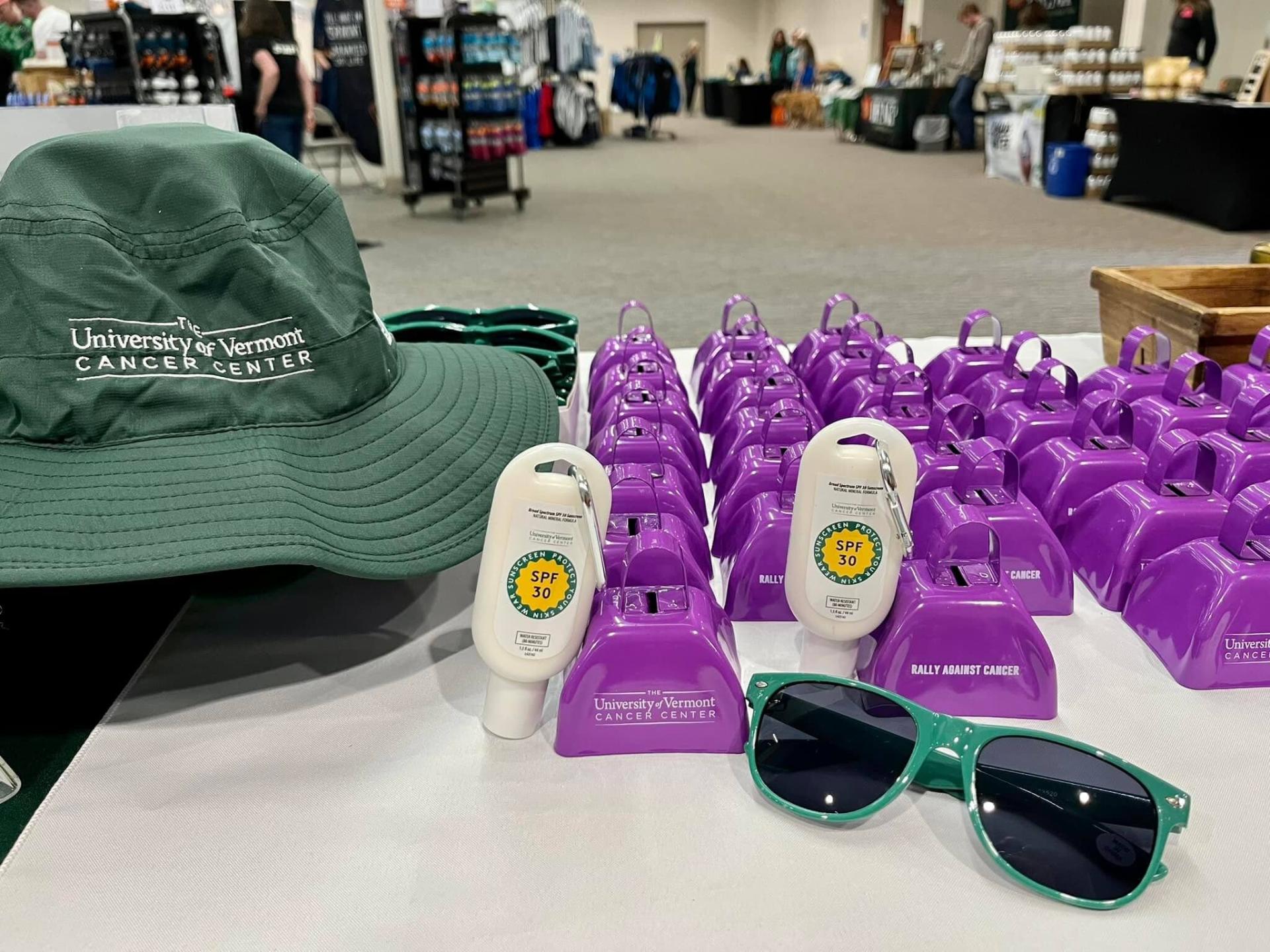 UVM Cancer Center branded sunscreen, pair of sunglasses, and some purple cowbells on a table