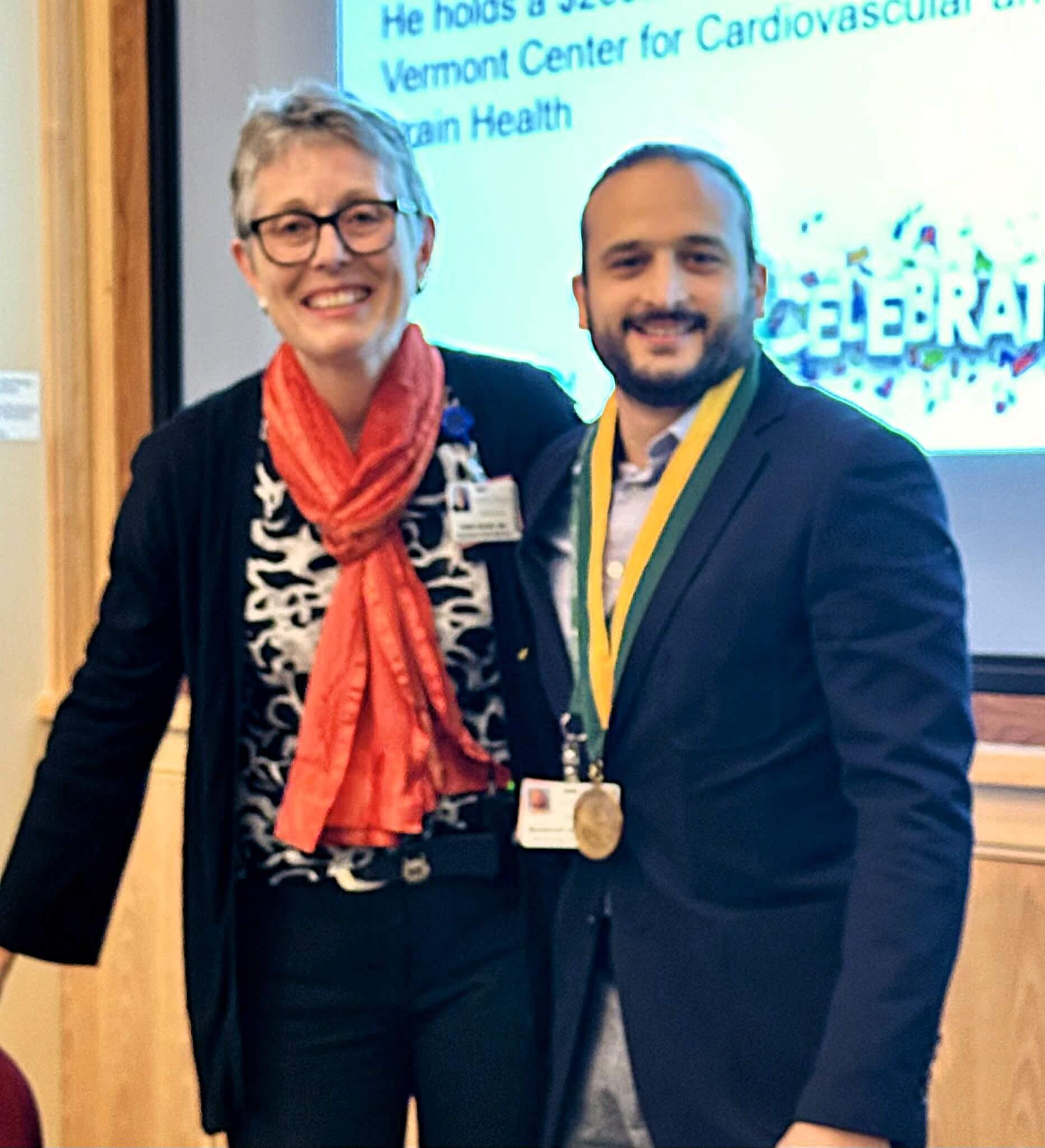Two researchers are posing together, smiling, with one of them adorned with an award while posing indoors.