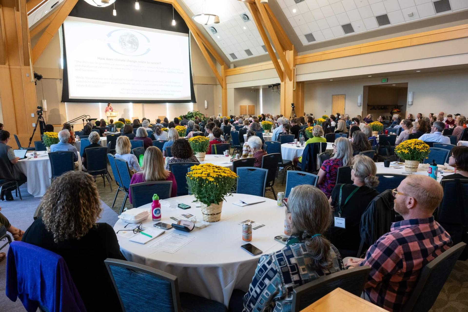 Room full of people sitting at round tables, watching a presentation
