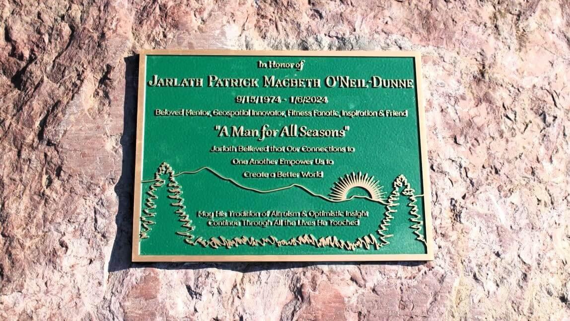 The memorial plaque for Jarlath O'Neil-Dunne placed outside the Aiken Center at UVM - the plaque is green and gold and depicts an enscription, trees, mountains, and the sun.