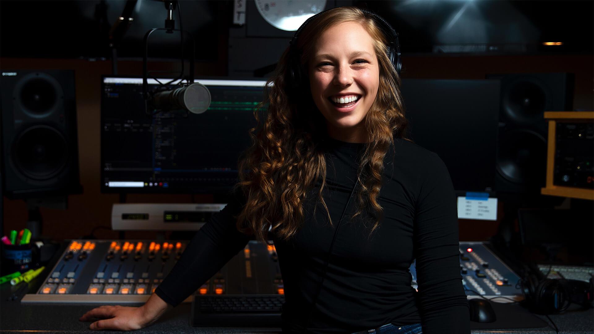 smiling woman with long hair and black top standing in front of sound recording equipment