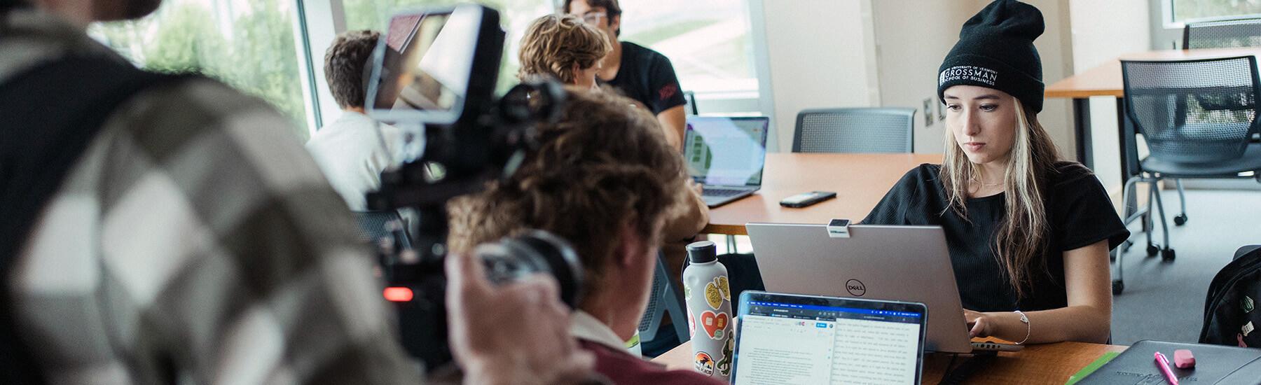 A woman focuses on her laptop while a another person films her