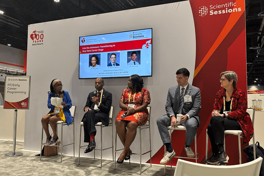 Five people are sitting together in a panel discussion indoors, in front of a backdrop with a TV.