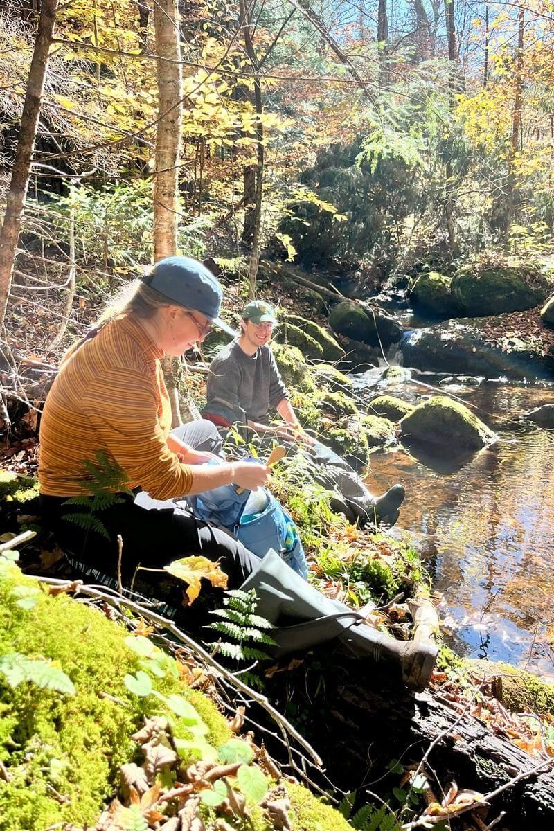 Two students sit by a stream to eat food and record information