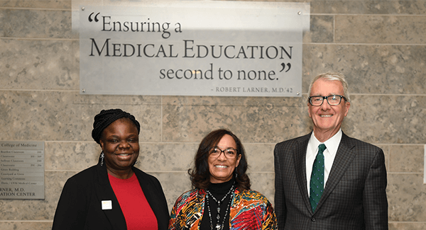 three people standing against a wall with a plaque saying "Ensuring a medical education second to none"