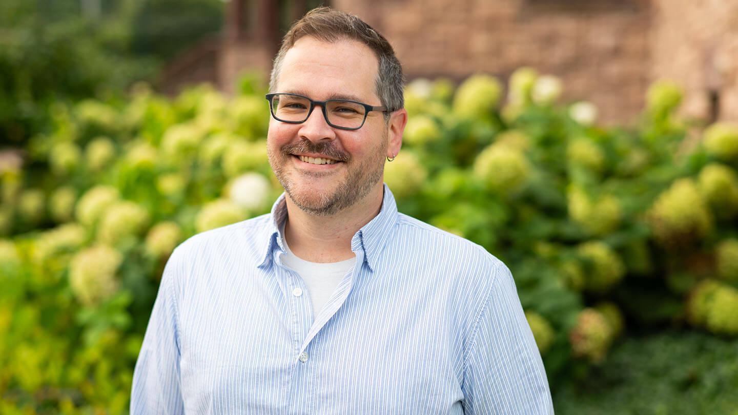 Cody Silfies, a young man in glasses and a button down shirt, smiles while standing outside. Behind him are white blossoms.