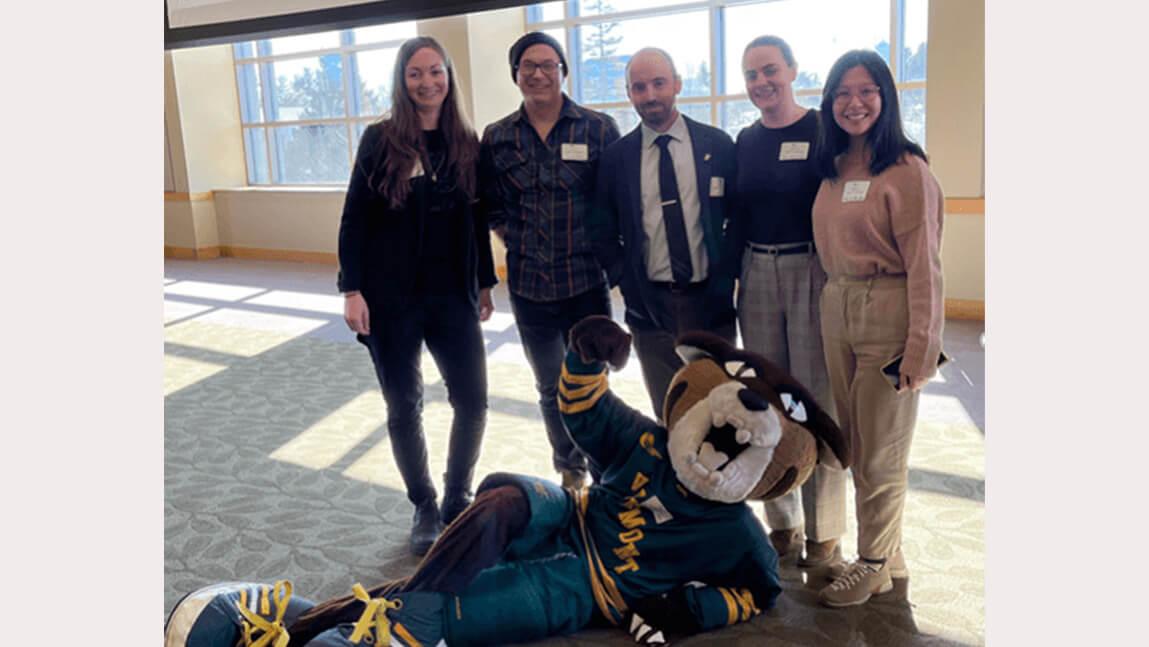 Five individuals stand in a line with the Rally Cat mascot laid on the floor in front of them in a pose.