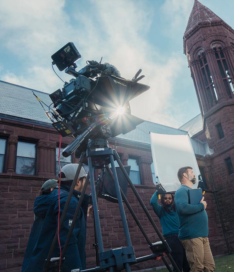 A group of people gathered around a camera in front of a building, engaged in conversation