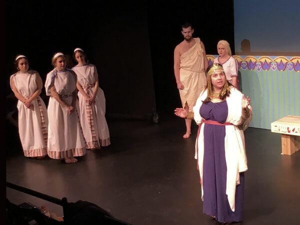  A woman in a dress stands on stage alongside several other individuals, all engaged in a presentation or performance