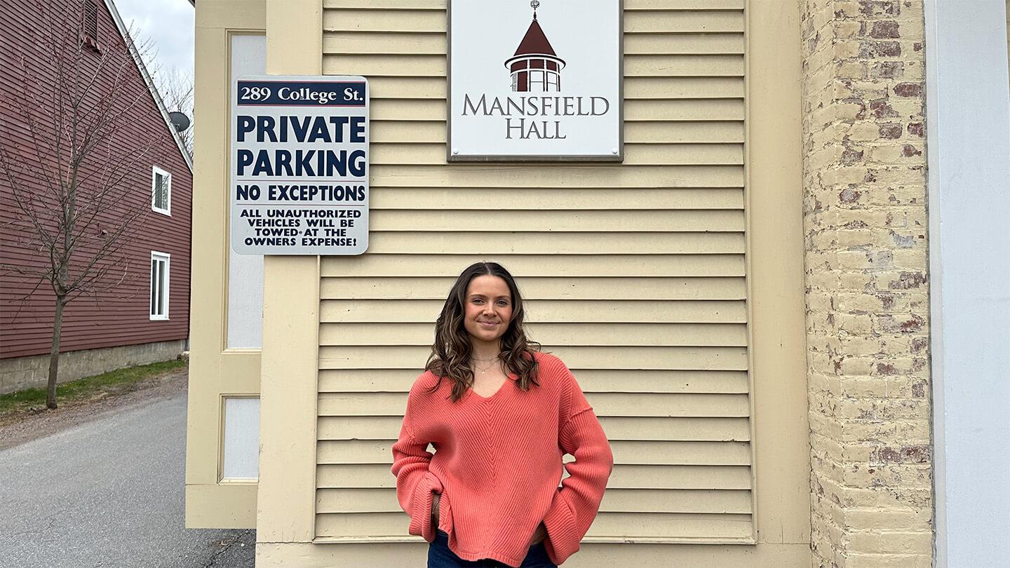 Young woman with long brown hair and orange sweater standing in front of yellow building with "Mansfield Hall" sign