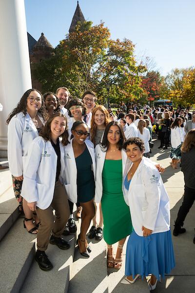 A group of smiling people standing outside on the steps of a building on a sunny day, wearing white coats.