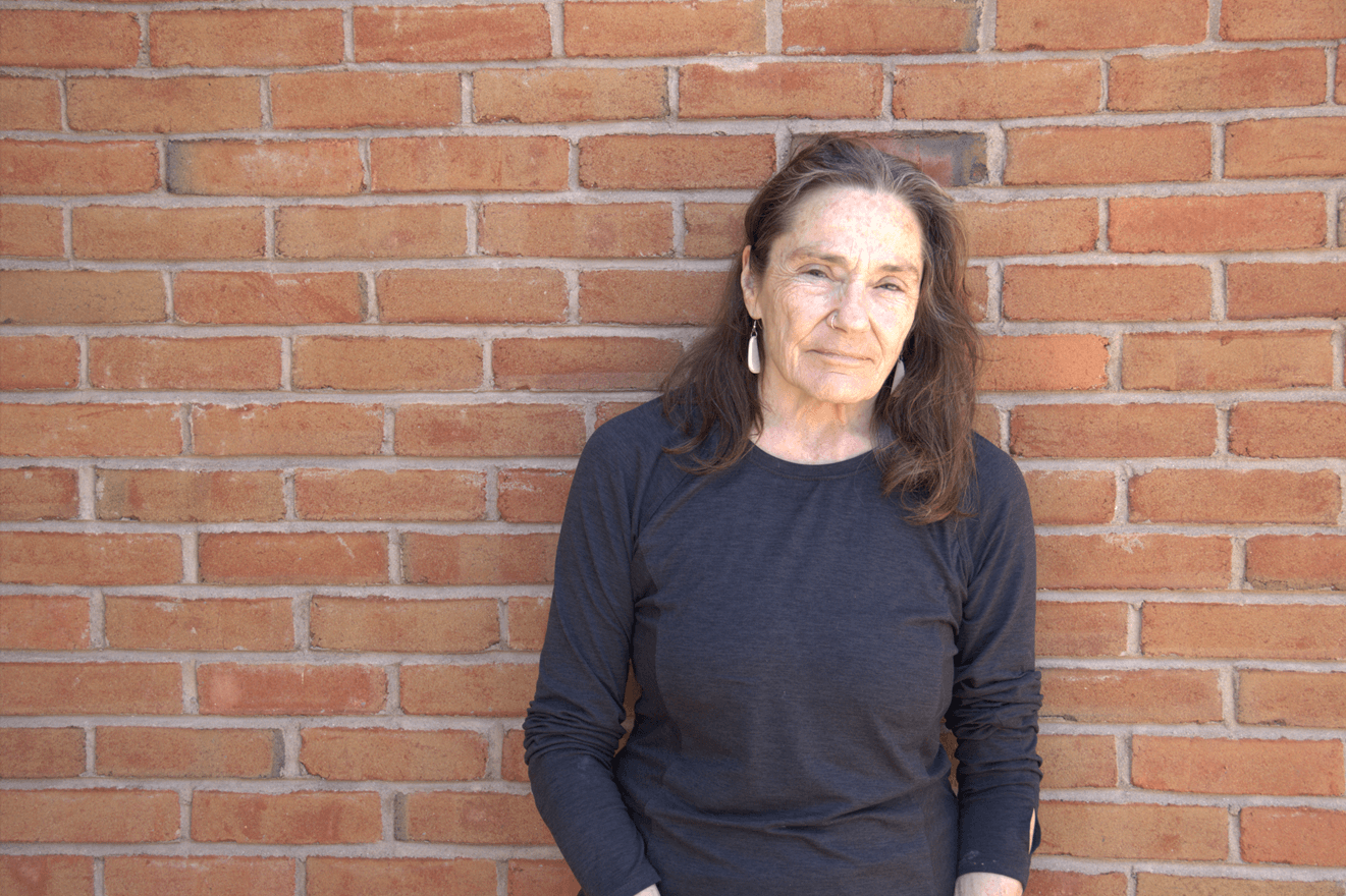 A woman with brown hair wearing a black shirt stands in front of a brick wall. 