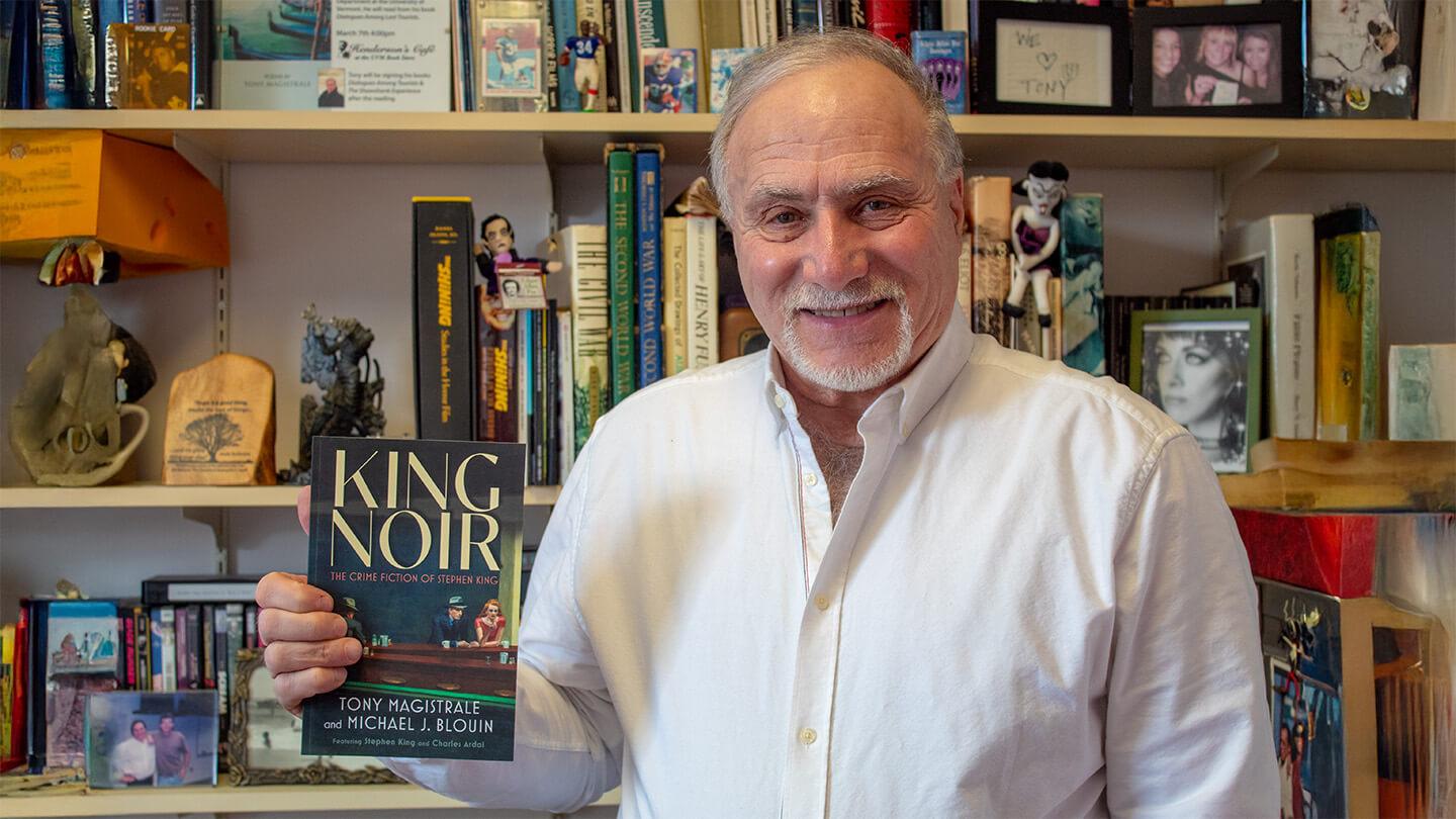 Professor Tony Magistrale, a man wearing white shirt with white hair and beard, holding "King Noir" book while standing in front of bookshelf