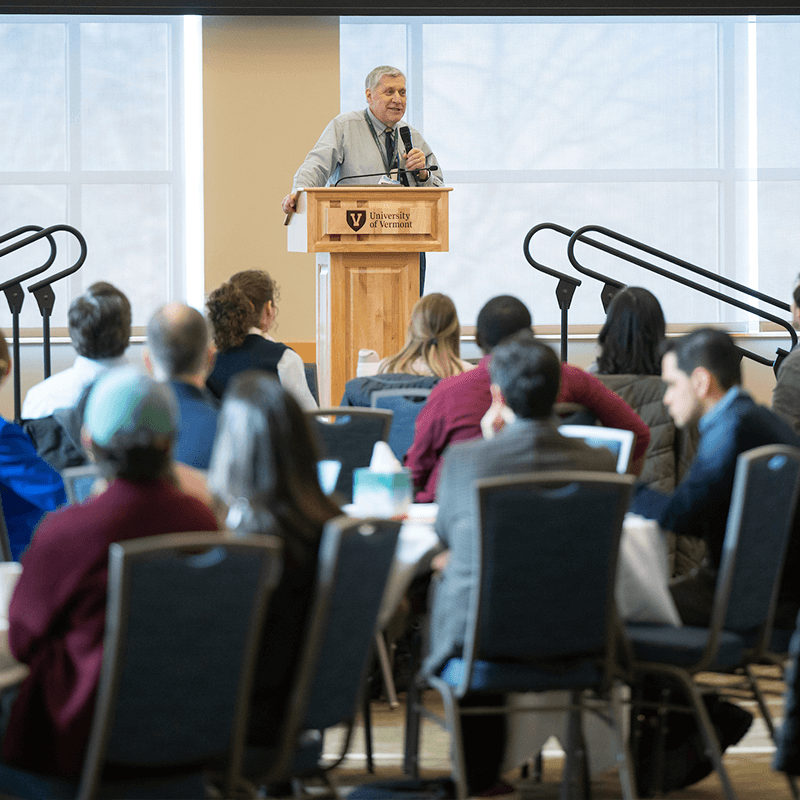 Dr. Holcombe standing at a podium in front of a small crowd of seated people.