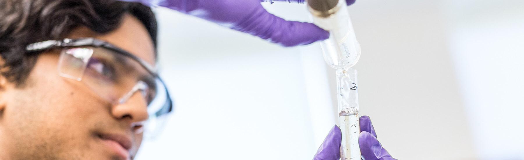 A person wearing a lab coat and purple gloves is holding a test tube