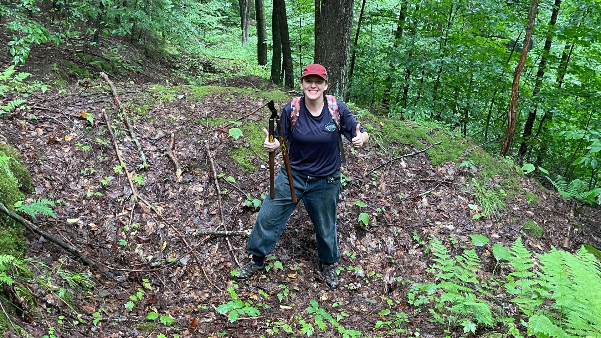 Es Sweeney standing in a forest holding large gardening shears and doing a thumbs up.