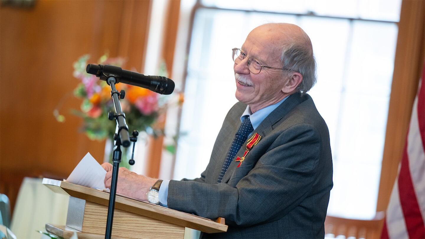 Wolfgang Mieder speaks at the podium in Memorial Lounge while wearing Order of Merit on lapel, microphone in foreground