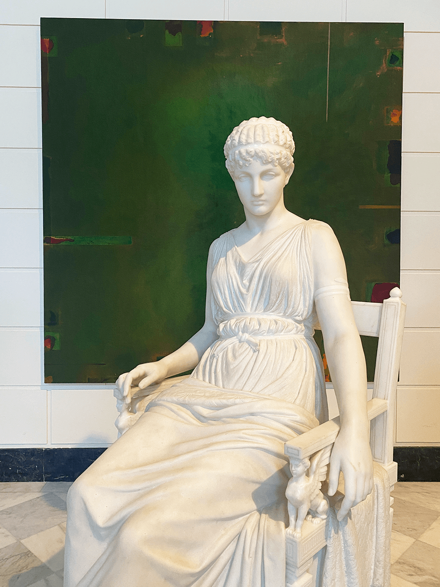 Cream marble statue of a seated woman, Penelope, in front of a large, square painting with layers of green.