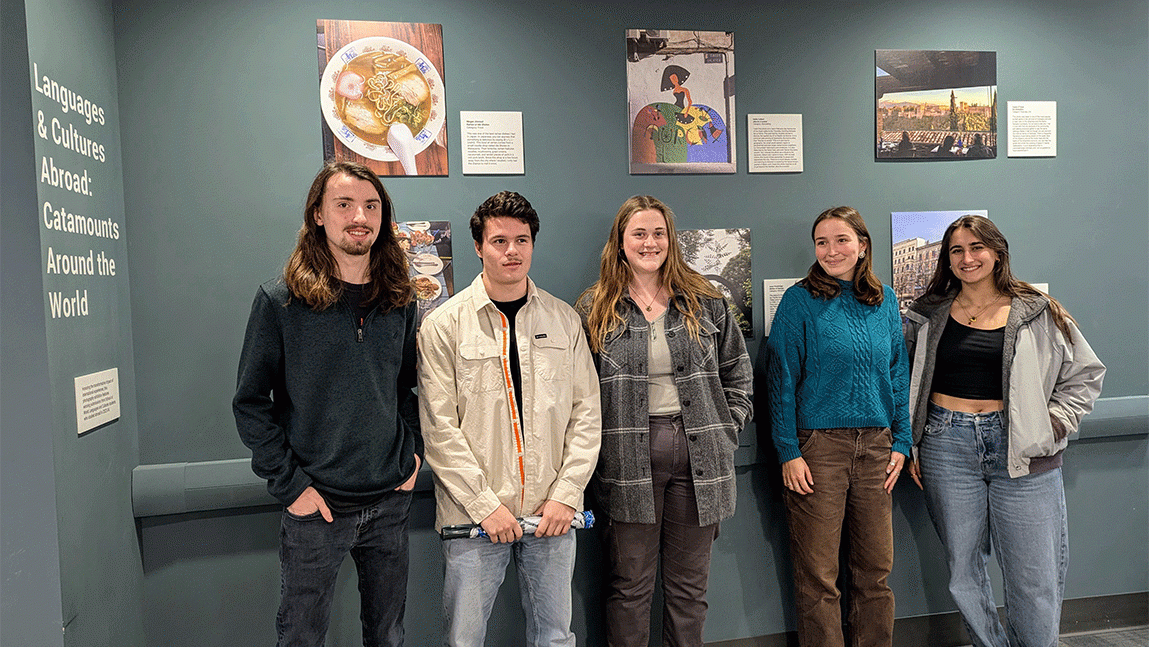 Five students stand in a row in front of a gallery of six photos. To the left of the students, wall text reads "Languages and cultures abroad: catamounts around the world."