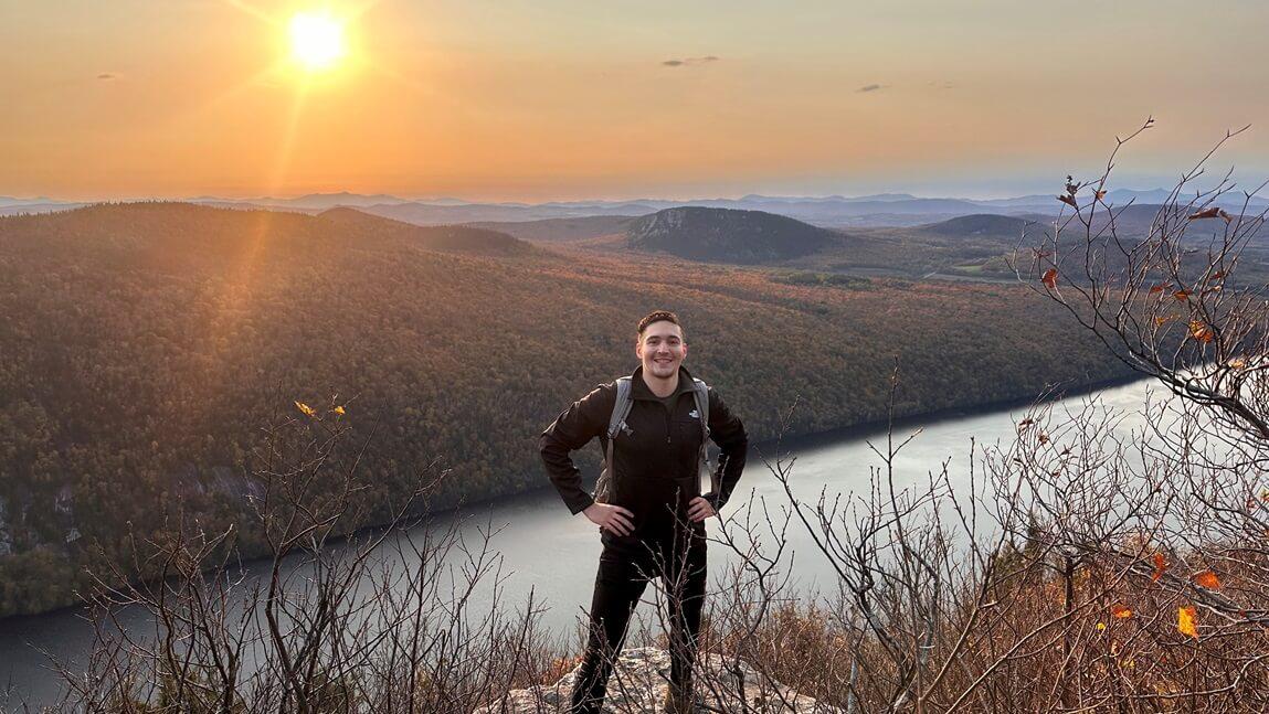 Rigo Melgar standing at a mountainous lookout with the sunsetting behind him and a lake below