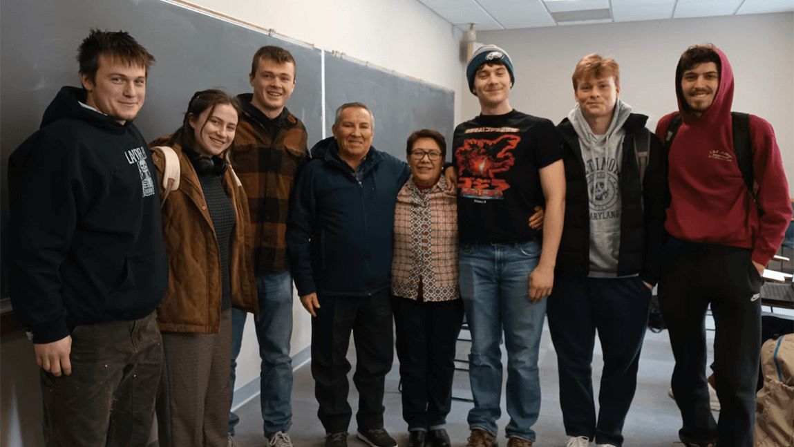 Students standing in a group indoors with a blackboard behind them. 