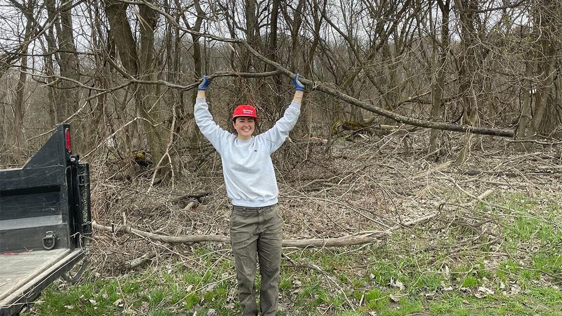 Young woman in grey sweatshirt and red hat smiling while holding a small branch overhead