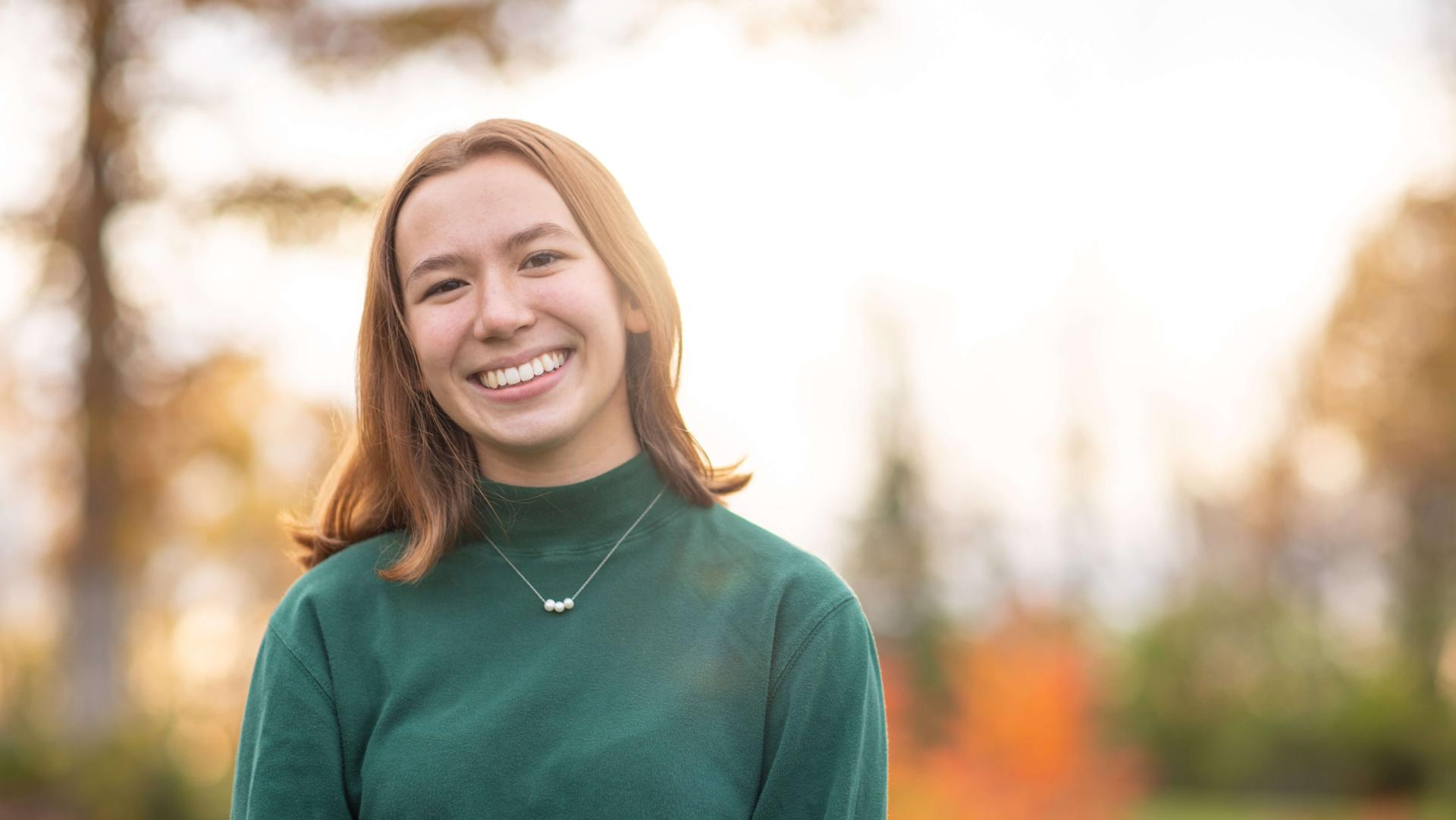 young woman in green turtleneck with shoulder-length brown hair smiling and standing in front of blurred forest background