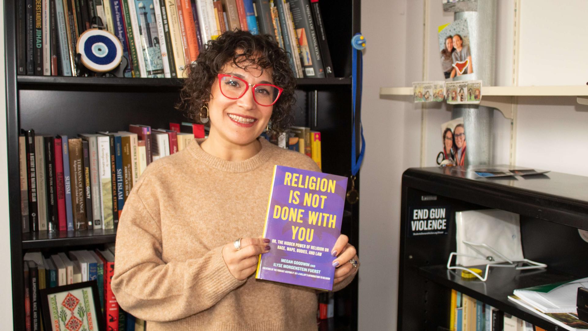 woman in beige sweater with curly brown hair and glasses standing in front of bookshelf holding a book