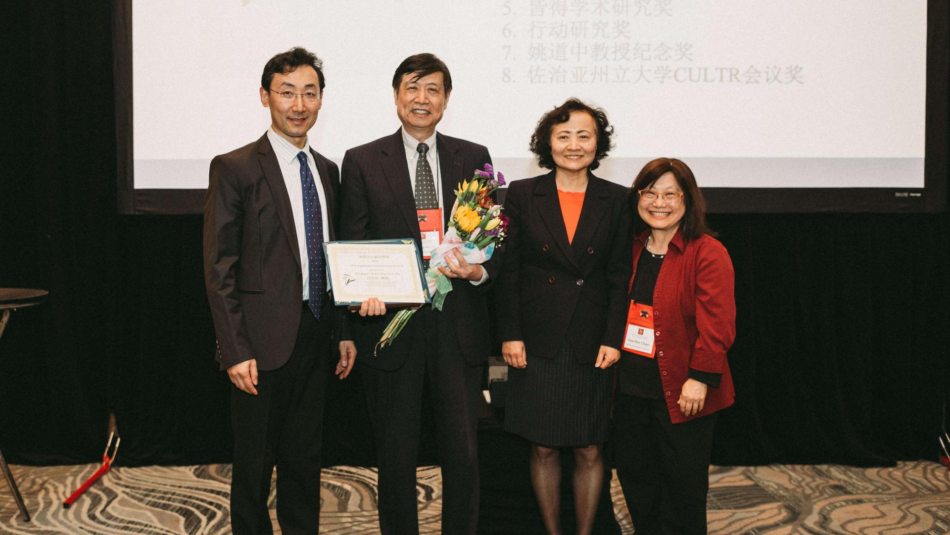 Four people standing in a line at a conference; man second from left holds an award he has received