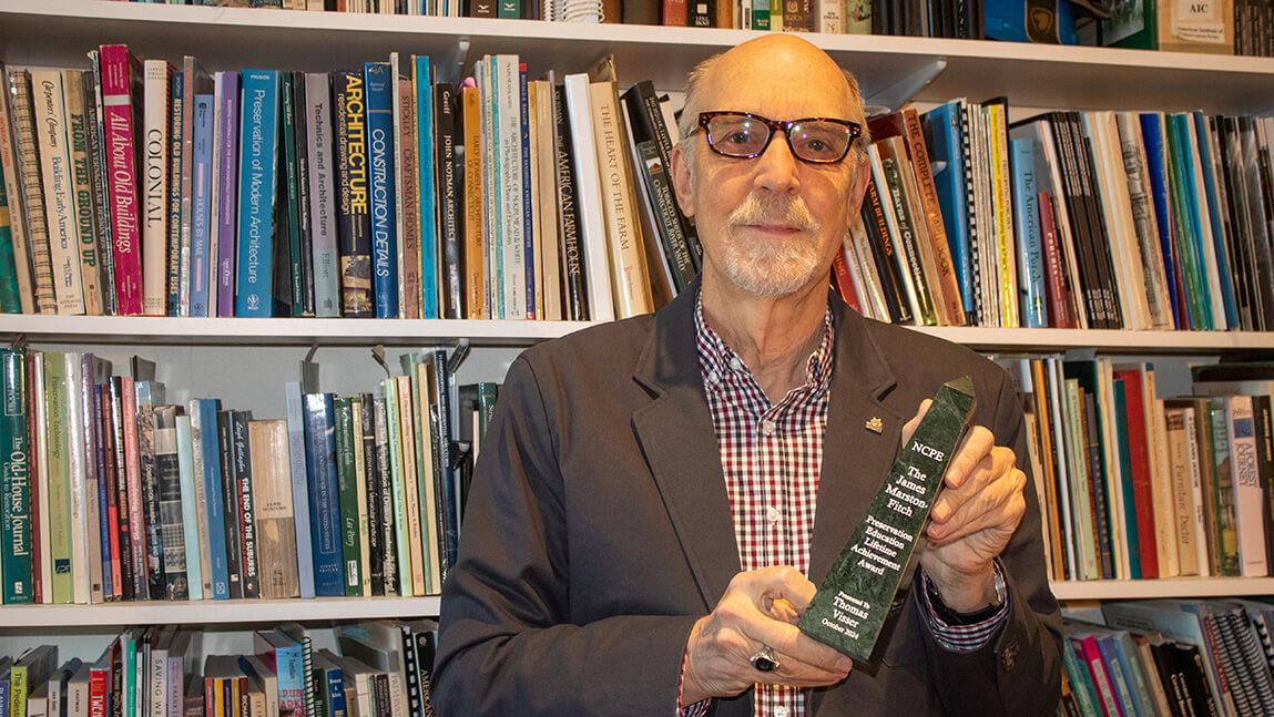 older male professor with glasses and brown suit jacket holding award while standing in front of book case in office