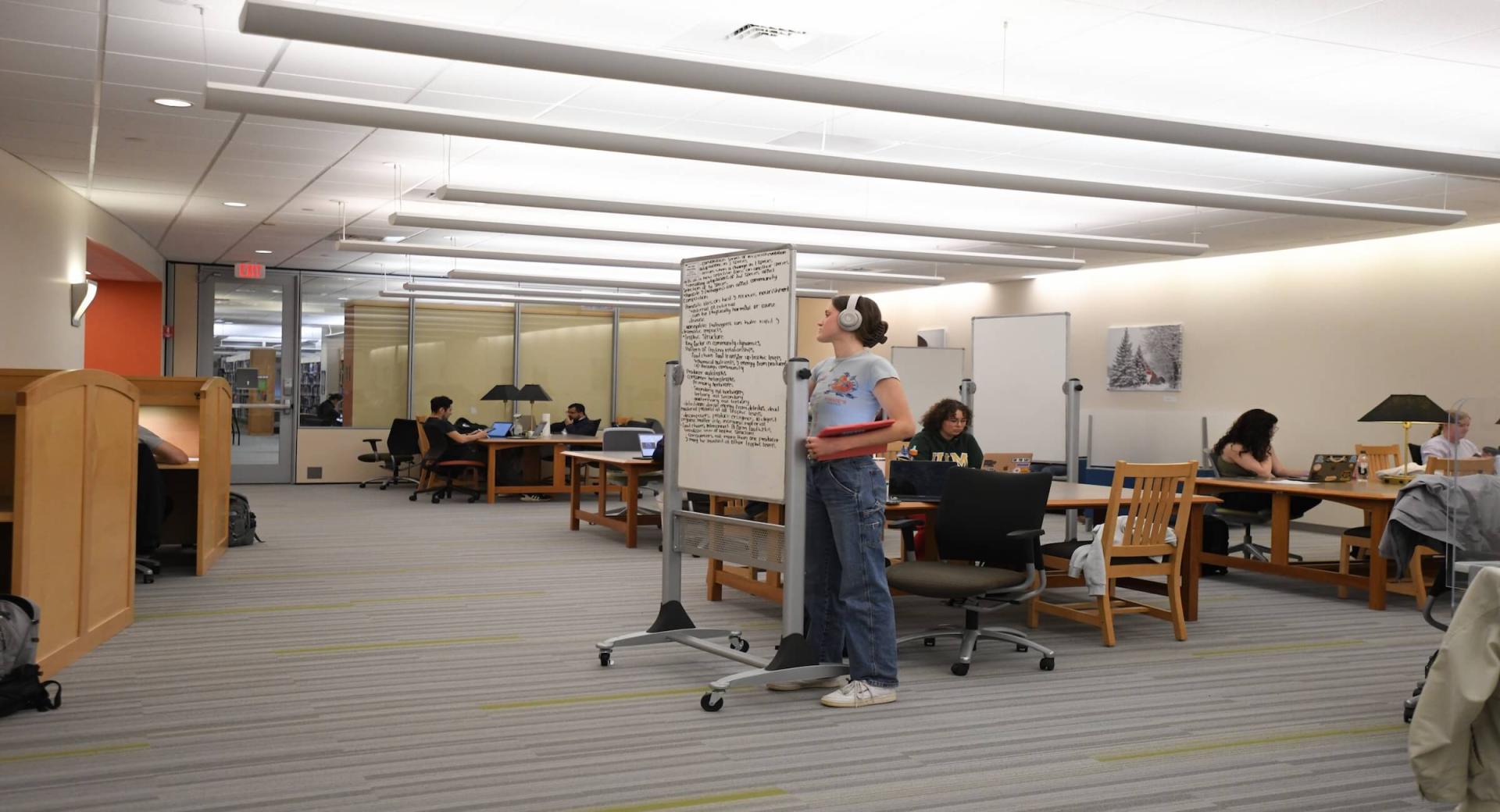 In Dana Health Sciences Library, a student is writing on a big white board while other students are sitting at tables and studying behind her. 