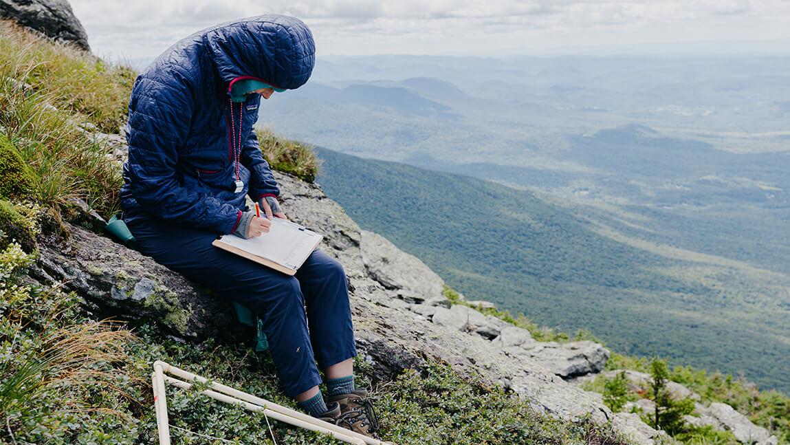 A researcher sitting on a mountain side with a clipboard