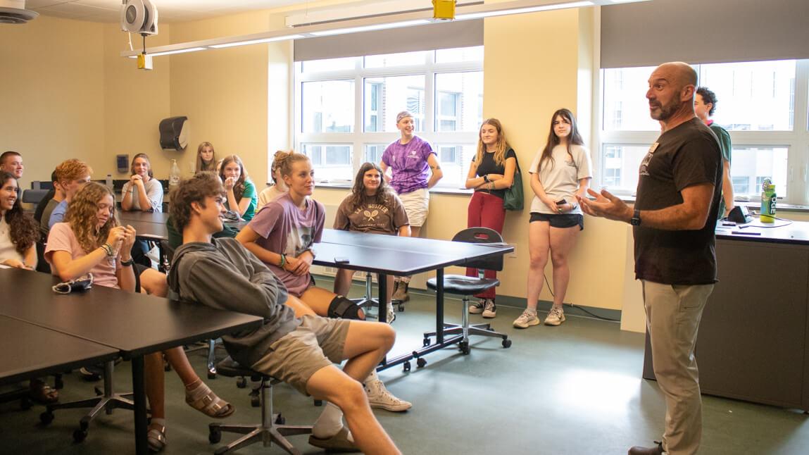 Peter standing in the front of a classroom addressing students. Some of the students are sitting, others are standing against the wall.