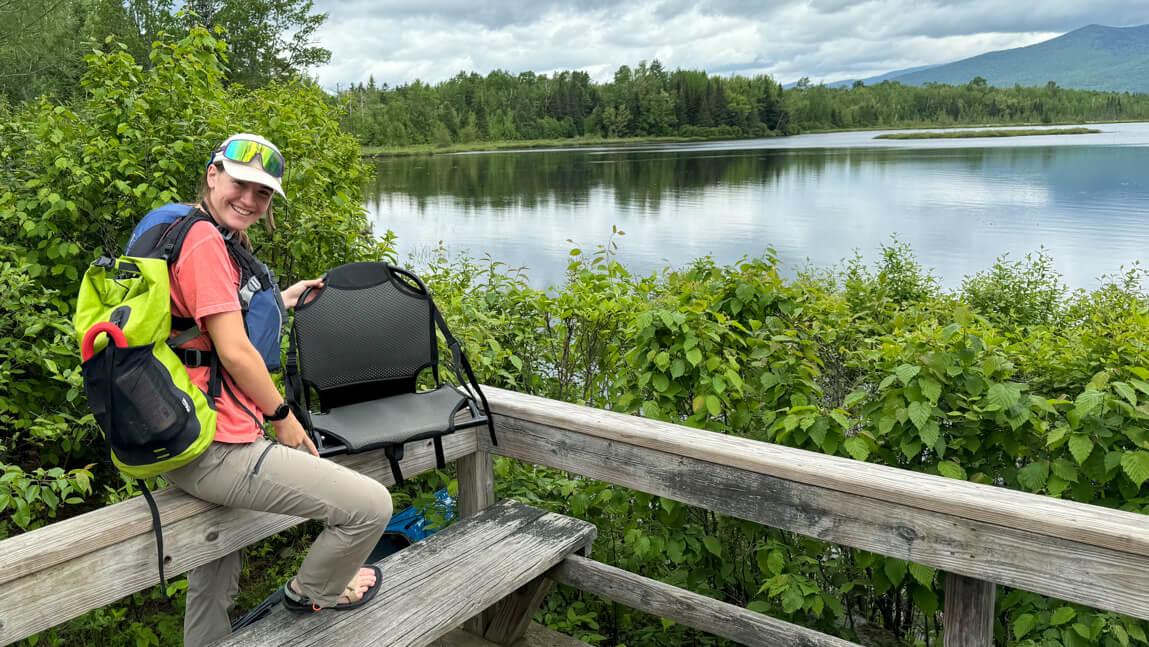 Lauren sitting on a wooden railing holding a camp chair and wearing a green backpack. Lake, mountain, and forest in the background.
