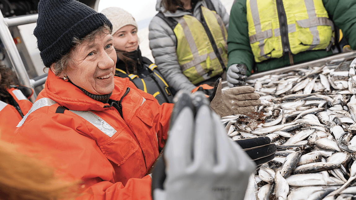 a woman in an organge coat stands in front a pile of fish and holds one up to the camera